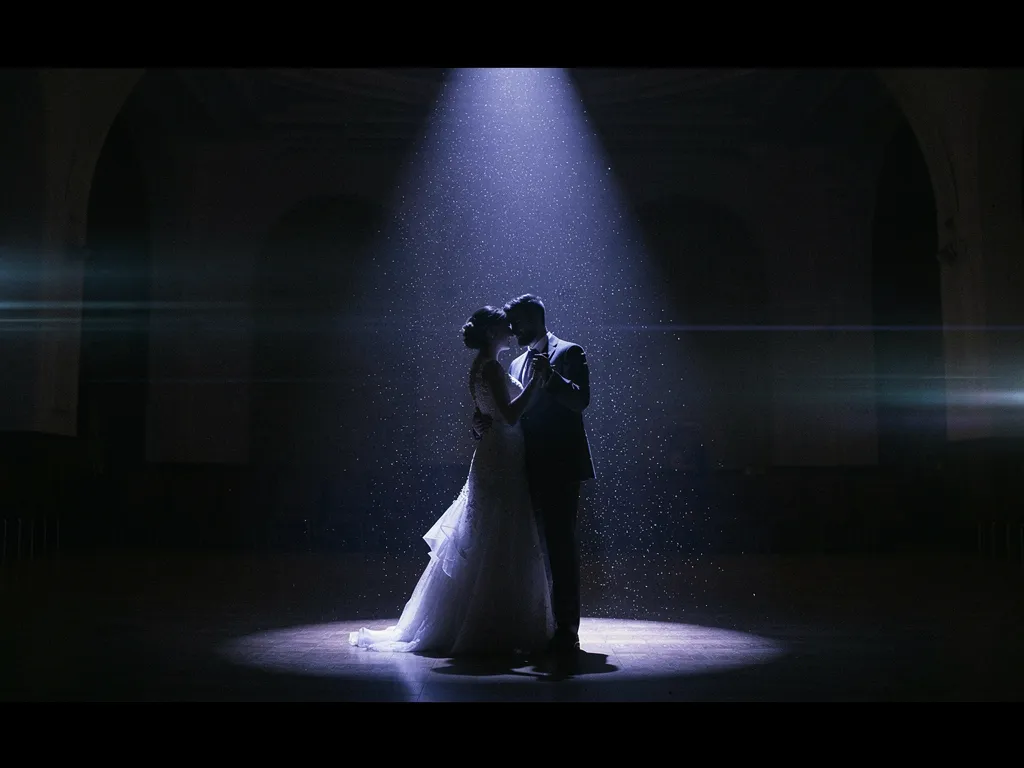 A bride and groom dancing alone under a single theatrical spotlight in a dark hall